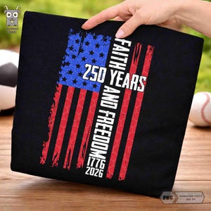 May include: Black t-shirt featuring a distressed American flag design with blue stars and red stripes. The text reads "250 YEARS FAITH AND FREEDOM 1776-2026". The shirt is held up by a hand, with a baseball and soccer ball in the background.
