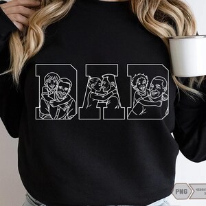 May include: Black sweatshirt with the word "DAD" in white block letters. Each letter features a line drawing of a father with his child. The sweatshirt is a crew neck style and has long sleeves. A white mug is held in the hand.
