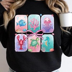 May include: A black sweatshirt with a colorful graphic of sea creatures in a gift box design. The graphic features a pink bow, a blue fish, a pink jellyfish, a red lobster, a green sea turtle, and a blue seahorse.