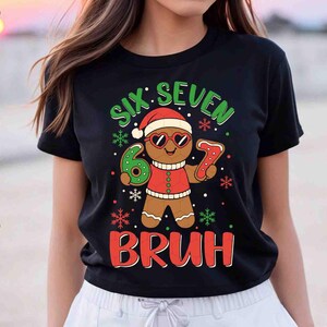 May include: Black t-shirt with a gingerbread man graphic wearing sunglasses and a Santa hat. The text reads "SIX SEVEN BRUH" in green and red. The gingerbread man is holding a number 6 and a number 7.