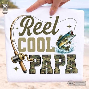 May include: White t-shirt with a fishing theme. The design features the words "Reel Cool Papa" in olive green and camouflage, a fishing rod, and a leaping fish with water splashes.