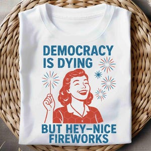 May include: White t-shirt with the text "DEMOCRACY IS DYING BUT HEY-NICE FIREWORKS" in blue. The design includes a red illustration of a woman holding a sparkler and red and blue fireworks.