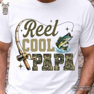 May include: White t-shirt with a fishing-themed graphic. The design includes a fishing rod, a jumping fish, and the words "Reel Cool Papa" in a combination of olive green and camouflage lettering. The shirt is a casual wear item.