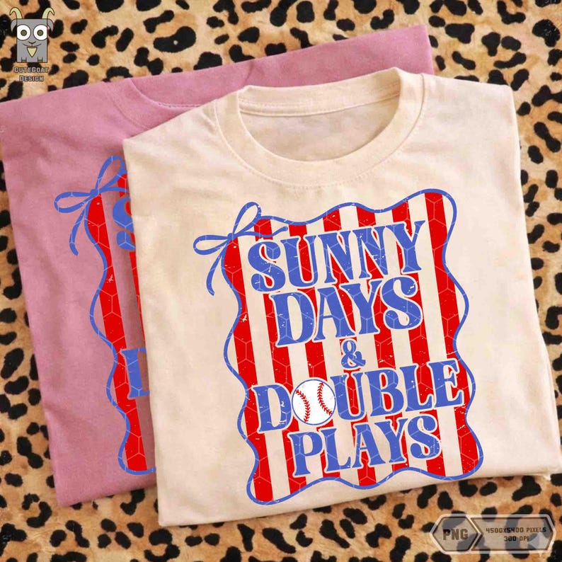 Puede incluir: Se muestran dos camisetas, una rosa claro y otra color crema. La camiseta crema presenta el texto "SUNNY DAYS & DOUBLE PLAYS" en azul y rojo, con un gr&aacute;fico de b&eacute;isbol. Las camisetas est&aacute;n dobladas sobre una superficie con estampado de leopardo.