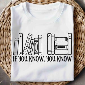 May include: White t-shirt featuring a black line drawing of a bookshelf with various book designs. The text "IF YOU KNOW, YOU KNOW" is printed below the books. The shirt is displayed on a woven surface.