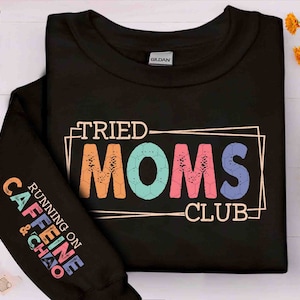 Pode incluir: Camisola de moletom preta com o texto "Tried Moms Club" em uma fonte colorida e desgastada. O texto "Running on Caffeine & Chaos" é impresso na manga em um esquema de cores arco-íris.