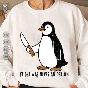 May include: A white crewneck sweatshirt featuring a graphic of a black and white penguin holding a kitchen knife. The penguin has orange feet and a beak. Below the graphic, the text reads, "FLIGHT WAS NEVER AN OPTION" in black capital letters.
