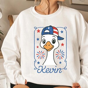 May include: White sweatshirt with a cartoon goose wearing a red, white, and blue baseball cap. The goose is wearing a red, white, and blue baseball cap with stars. The text "Kevin" is printed below the goose.