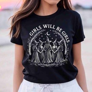 May include: Black t-shirt with the text "GIRLS WILL BE GIRLS" in white. The graphic features five witches holding hands under a crescent moon and stars. The witches are wearing pointed hats and long dresses.