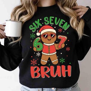 May include: Black sweatshirt with a gingerbread man graphic wearing heart-shaped sunglasses and a Santa hat. The text reads "SIX SEVEN BRUH" in green and red, with snowflakes. The gingerbread man is holding a number 6 and a number 7.
