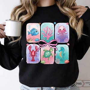May include: Black sweatshirt with a colorful graphic of a gift box filled with sea creatures. The box is tied with a pink ribbon and contains a seahorse, a turtle, a lobster, a jellyfish, and a fish.