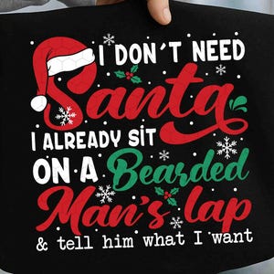 Op de afbeelding: Zwart grafisch ontwerp met de tekst "I DON'T NEED Santa, I ALREADY SIT ON A Bearded Man's lap & tell him what I want." Het ontwerp bevat een rode kerstmuts, sneeuwvlokken en hulst.