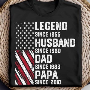 Puede incluir: Camiseta negra con un dise&ntilde;o de bandera americana desgastada. La camiseta presenta las palabras "LEGEND SINCE 1955, HUSBAND SINCE 1980, DAD SINCE 1983, PAPA SINCE 2010" en letras blancas. La camiseta se muestra sobre una superficie tejida.