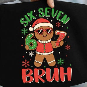 May include: Black graphic tee featuring a gingerbread man wearing heart-shaped sunglasses and a Santa hat. The text reads "SIX SEVEN BRUH" in green and red, with snowflake accents. The gingerbread man is holding a number 6 and a number 7.