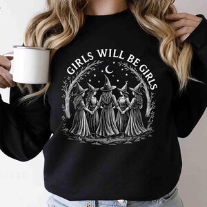 May include: Black sweatshirt with a white graphic of five witches holding hands, surrounded by a crescent moon, stars, and the text "GIRLS WILL BE GIRLS". A white mug is held in the left hand.