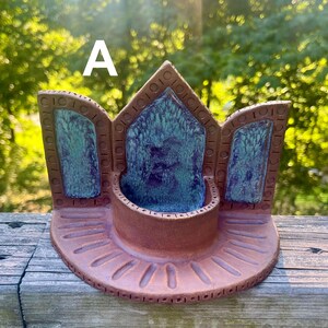 May include: A terracotta ceramic incense holder featuring a central circular basin and three pointed back panels. The panels and basin interior are glazed in a mottled teal and deep purple finish. The base has etched radial lines and the letter A.