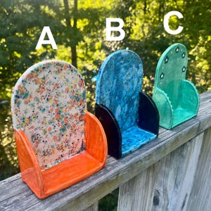 May include: Three ceramic wall pockets labeled A, B, and C. A is white with multicolored speckles and an orange base. B is blue with a mottled texture and a black base. C is light green with small flower patterns and a matching green base.