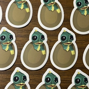 May include: A set of ten stickers featuring a cartoon green and blue dinosaur hatching from a brown egg. The dinosaur has large eyes and a toothy grin.