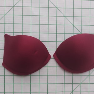 May include: Two burgundy bra cups, likely for a DIY project or replacement. The cups are a solid, deep red color and have a smooth, seamless appearance. They are placed on a green and white grid background.