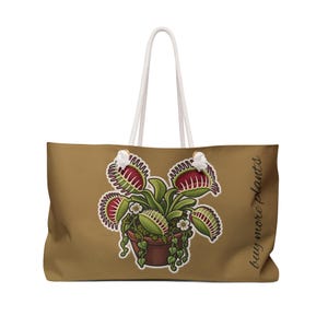 Può includere: Una borsa tote color marrone chiaro con manici in corda bianca, caratterizzata da un'illustrazione centrale di una pianta carnivora dionaea in vaso con foglie rosse e verdi e piccoli fiori bianchi, con il testo in corsivo buy more plants a destra.