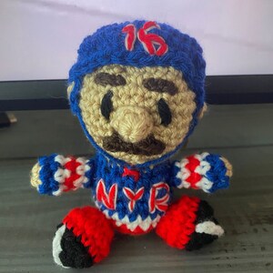May include: Crocheted toy figure of a hockey player wearing a blue and white jersey with the letters "NYR" and the number "16" on the hat. The figure has a brown mustache and eyebrows.