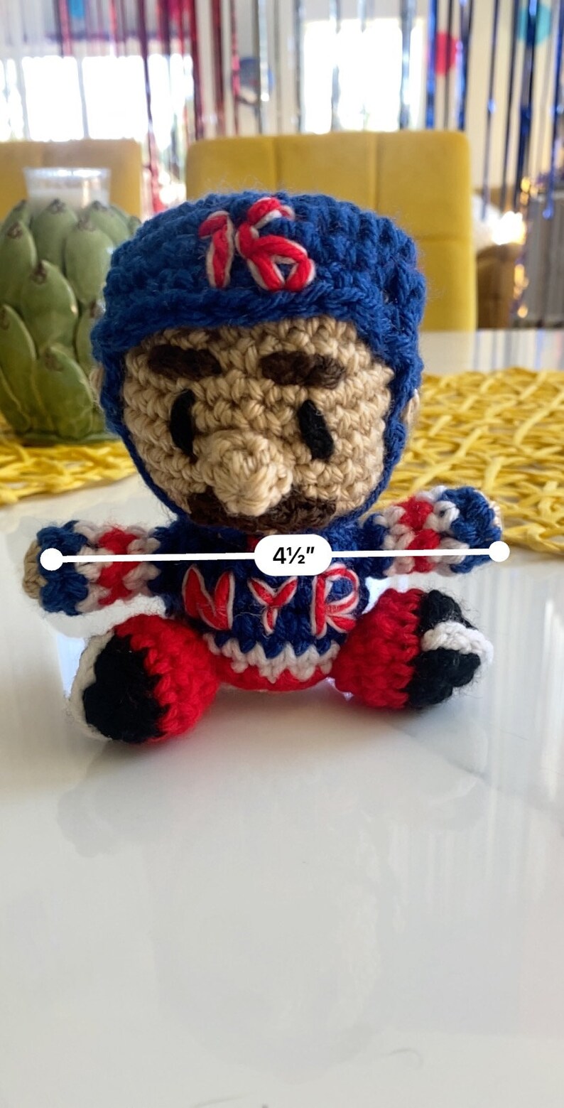 May include: Crocheted plush toy of a baseball player wearing a blue hat with the number 15 on it. The player is wearing a red, white, and blue jersey with the letters "NYR" on the front. The toy is 4 1/2 inches tall.
