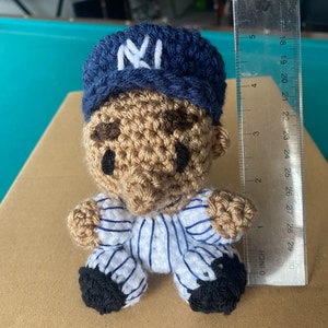 May include: A crocheted doll wearing a blue and white baseball uniform with a blue baseball cap with a white "NY" logo. The doll is sitting on a brown surface.