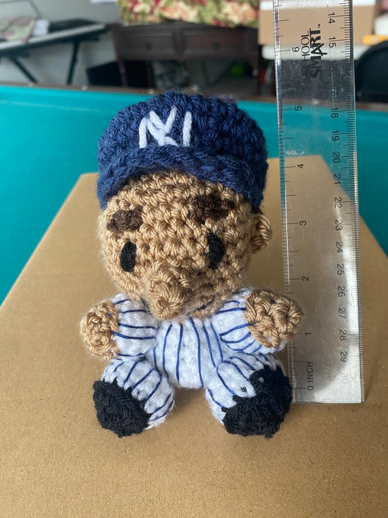 May include: A crocheted doll wearing a blue and white baseball uniform with a blue baseball cap with a white "NY" logo. The doll is sitting on a brown surface.