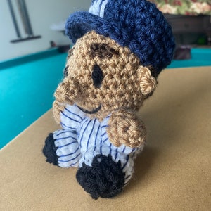 May include: A crocheted plush toy wearing a blue and white striped shirt and a blue hat. The toy has a brown face with black eyes and a smile.