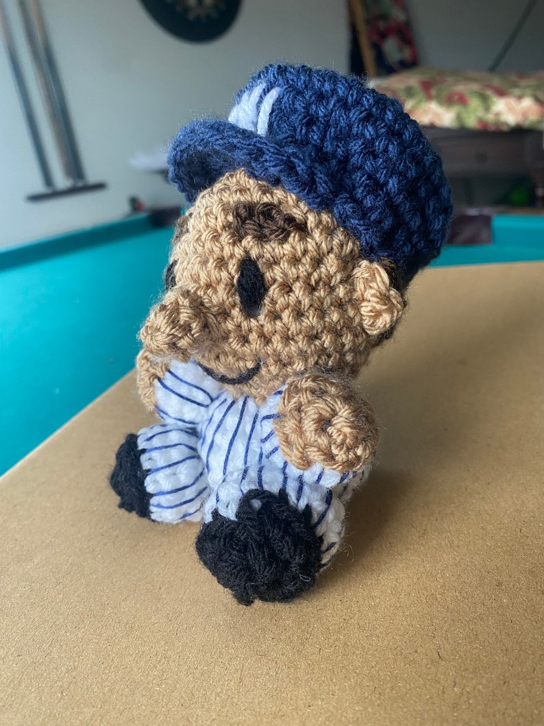 May include: A crocheted plush toy wearing a blue and white striped shirt and a blue hat. The toy has a brown face with black eyes and a smile.