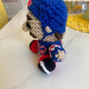 May include: A crocheted plush toy of a hockey player wearing a blue helmet, red and white jersey, and black and white skates.