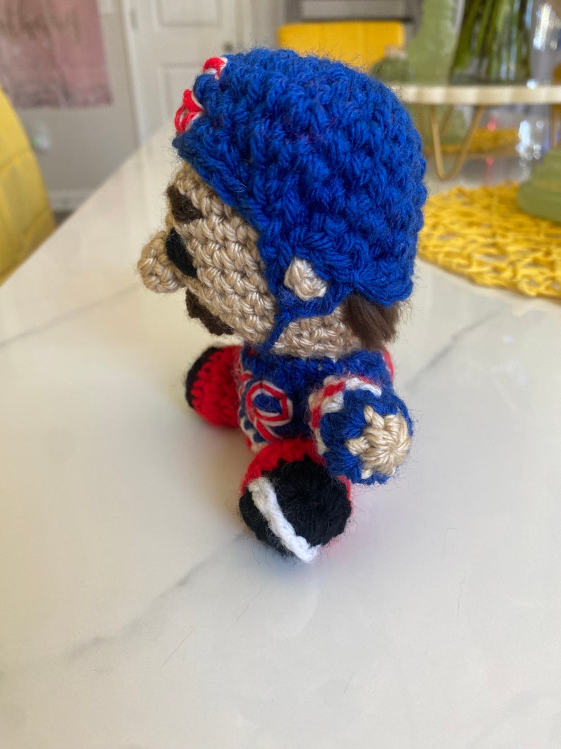 May include: A crocheted plush toy of a hockey player wearing a blue helmet, red and white jersey, and black and white skates.