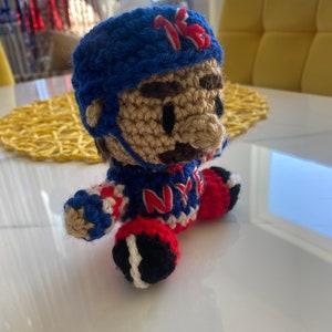 May include: Crocheted plush toy of a hockey player wearing a blue and white jersey with the letters "NY" on the front, a blue helmet with the number "16" on the side, and red and white pants. The toy is sitting on a white surface.