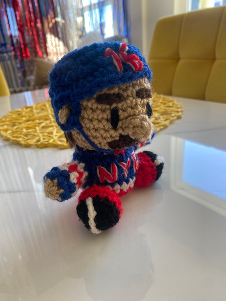 May include: Crocheted plush toy of a hockey player wearing a blue and white jersey with the letters "NY" on the front, a blue helmet with the number "16" on the side, and red and white pants. The toy is sitting on a white surface.
