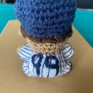 May include: A crocheted baseball player doll wearing a blue hat and a white and blue striped jersey with the number "99" on the back.