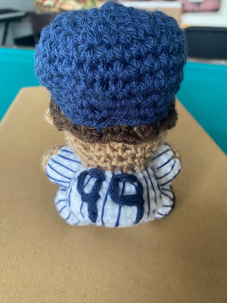 May include: A crocheted baseball player doll wearing a blue hat and a white and blue striped jersey with the number "99" on the back.