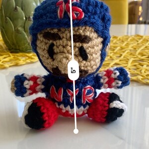 May include: A crocheted doll wearing a blue and red hockey jersey with the letters "NYR" on the front. The doll is 5 inches tall and has a brown face with black eyes.