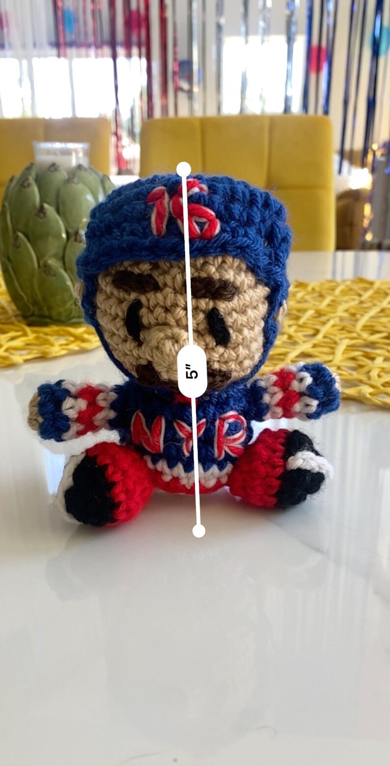 May include: A crocheted doll wearing a blue and red hockey jersey with the letters "NYR" on the front. The doll is 5 inches tall and has a brown face with black eyes.