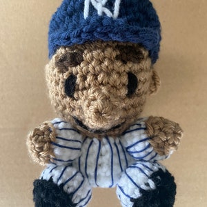 May include: A crocheted baseball player doll wearing a blue and white striped uniform with a blue baseball cap with a white "NY" logo.