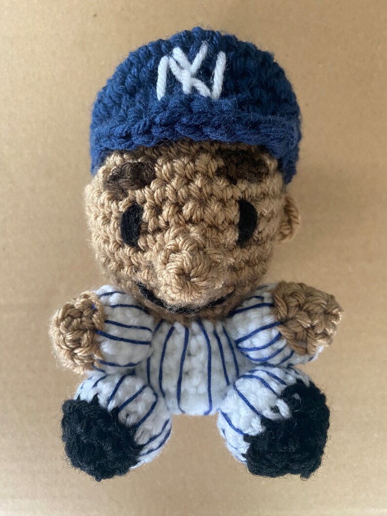 May include: A crocheted baseball player doll wearing a blue and white striped uniform with a blue baseball cap with a white "NY" logo.