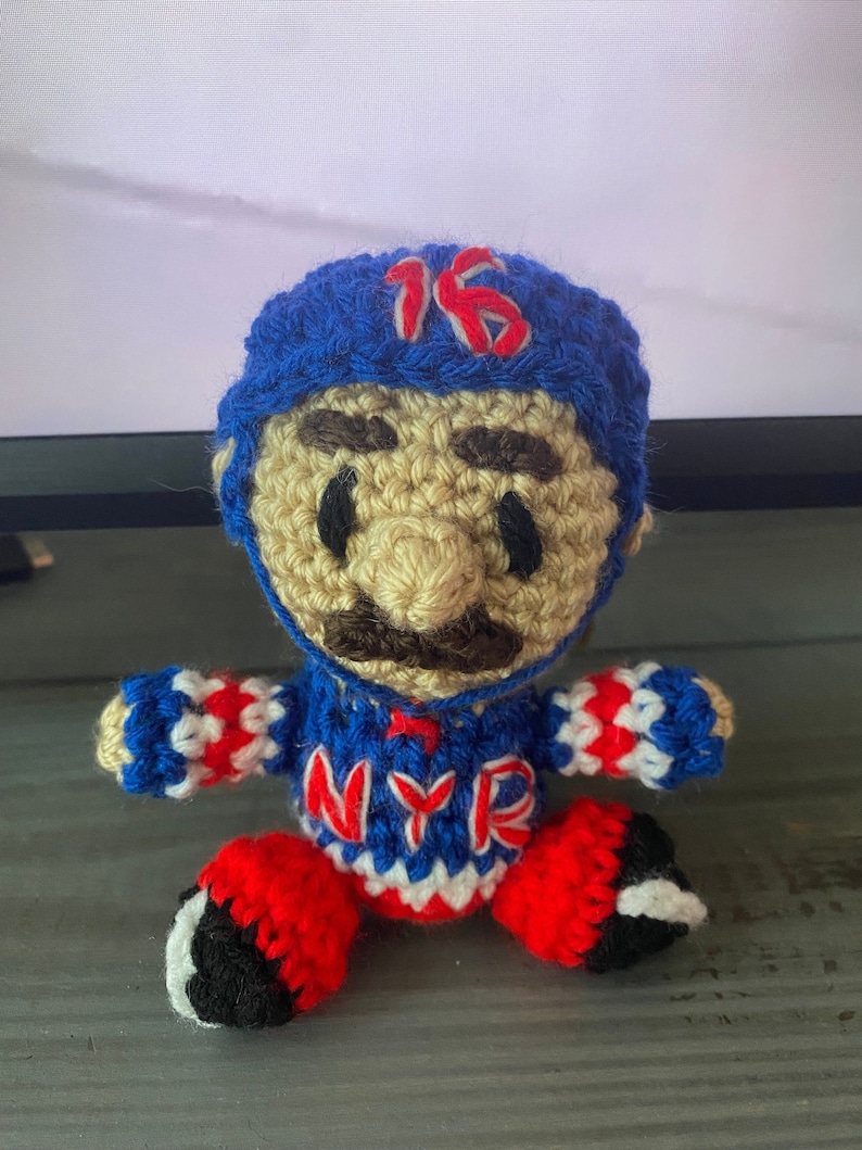 May include: Crocheted toy figure of a hockey player wearing a blue and white jersey with the letters "NYR" and the number "16" on the hat. The figure has a brown mustache and eyebrows.
