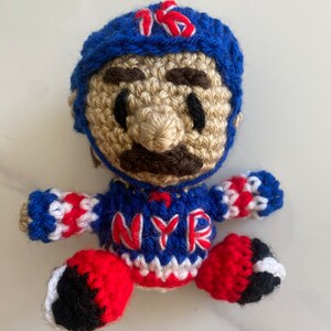 May include: Crocheted plush toy of a hockey player wearing a blue helmet with the number 16 on it, a red, white, and blue jersey with the letters NYR, and red and black pants.