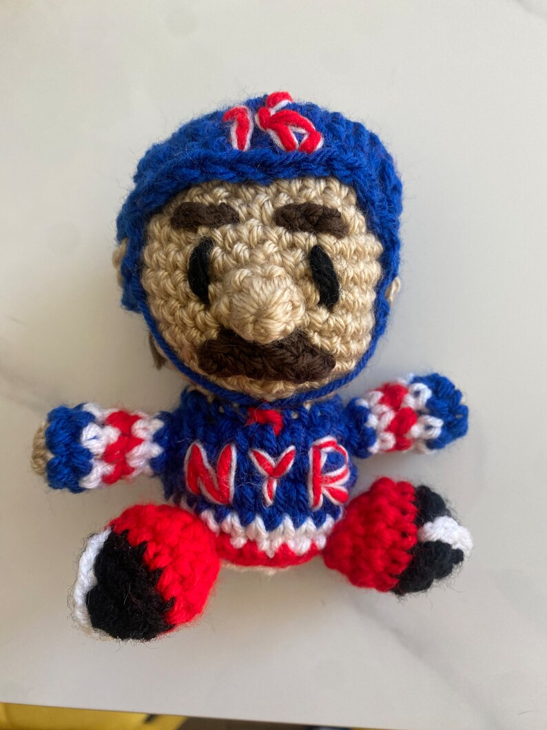May include: Crocheted plush toy of a hockey player wearing a blue helmet with the number 16 on it, a red, white, and blue jersey with the letters NYR, and red and black pants.