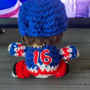 May include: Crocheted hockey player doll wearing a blue and white jersey with the number 16 on the back. The doll is wearing a blue hat and has brown hair.