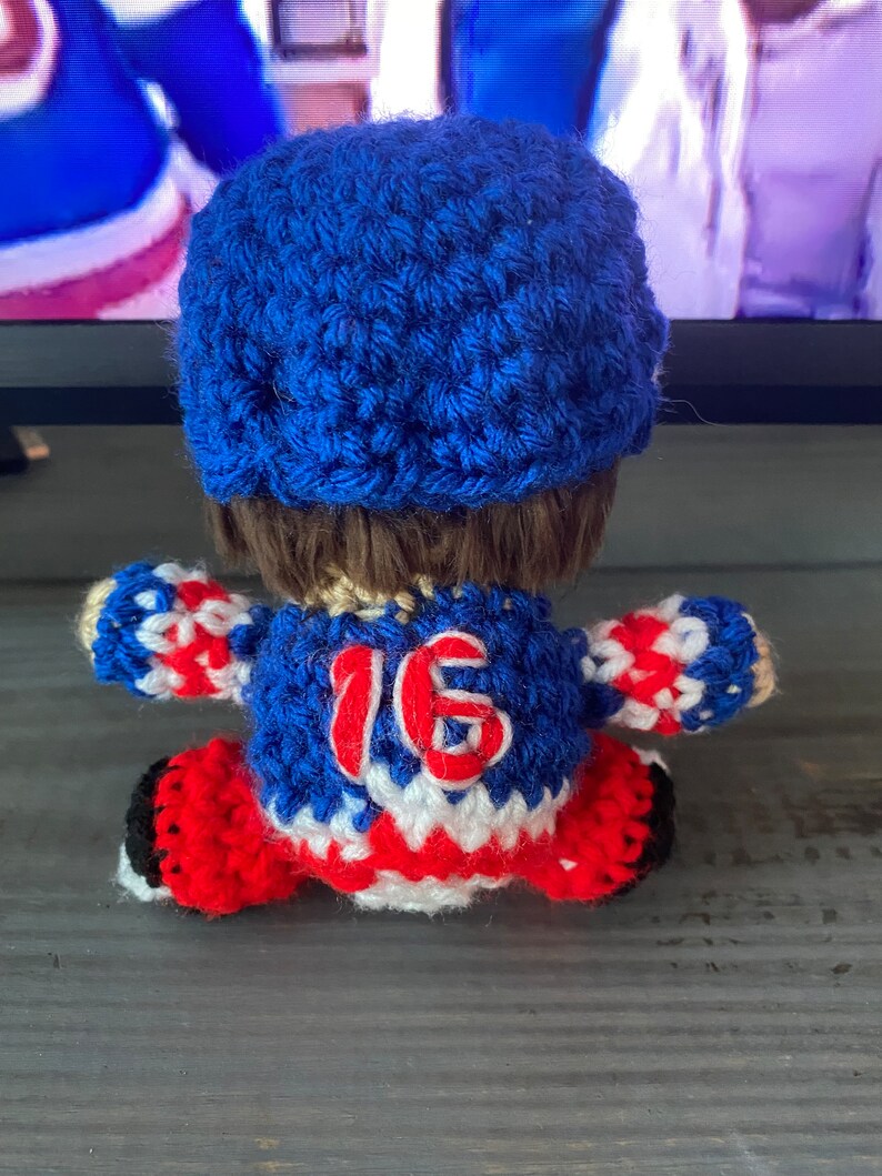 May include: Crocheted hockey player doll wearing a blue and white jersey with the number 16 on the back. The doll is wearing a blue hat and has brown hair.