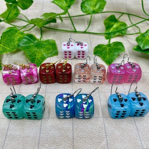 May include: A set of eight pairs of dangle earrings featuring dice with hearts and spades printed on them. The dice are made of clear resin with different colors, including pink, red, blue, and white.