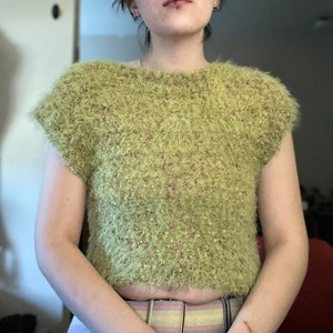 May include: A person wearing a green fuzzy cropped sweater with short sleeves and a belt. The person has short brown hair and is wearing a nose ring and lip ring.