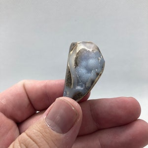 Op de afbeelding: Een kleine, lichtblauwe en bruine agaatgeode met een ruw, natuurlijk exterieur en een glad, gepolijst interieur. De geode is gevuld met kleine, witte kristallen.