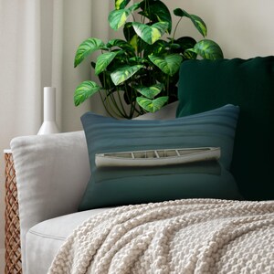 May include: A decorative pillow featuring a serene lake scene with a white canoe floating on the water. The pillow has a blue and teal color scheme, evoking a sense of tranquility. The pillow is placed on a gray sofa with a cream-colored throw blanket.