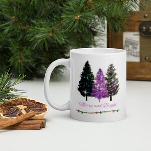 Op de afbeelding: Witte keramische mok met een ontwerp van drie kerstboom in tinten van paars, groen en zwart. De bomen zijn versierd met lichtjes en glitter. De tekst "Merry and Bright" is onder de bomen gedrukt met een reeks lichtjes.